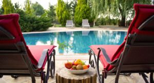 Backyard pool and chairs with fruit bowl in between, trees in background.